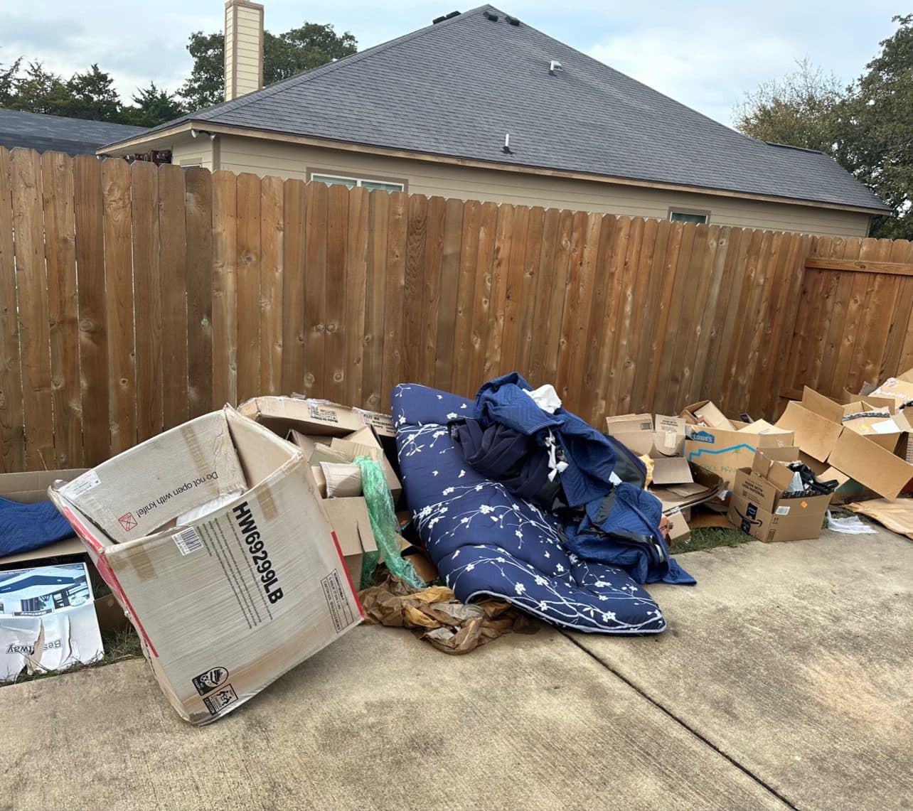 Backyard patio with boxes, a mattress, and debris piled against the fence before removal